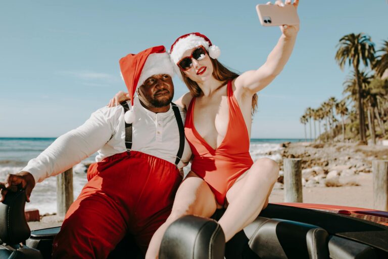 A couple dressed as Santa and an elf taking a selfie by the beach in summer.